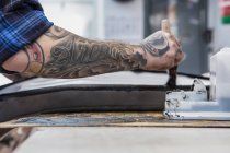 Crop side view of tattooed craftsman applying glue on motorcycle seat while making upholstery at workbench — Stock Photo