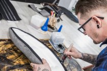 Da sopra vista laterale dell'artigiano tatuato che applica la colla sul sedile del motociclo mentre fa la tappezzeria al banco da lavoro — Foto stock