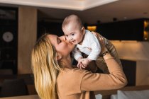 Seitenansicht der entzückten Mutter, die ihr entzückendes lächelndes Baby wirft und küsst, während sie zu Hause zusammen Spaß hat — Stockfoto