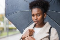 Young trendy African American female in warm coat standing with umbrella on modern city street and looking at camera — Stock Photo