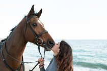 Seitenansicht der jungen Frau küsst Maulkorb von Fuchshengst in Zaumzeug gegen welligen Ozean — Stockfoto
