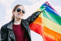 Знизу радісна лесбіянка стоїть на вулиці з ЛГБТ райдужним прапором, що пурхає вітром і озирається на хмарне небо. — стокове фото