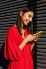 Молодий контент жінки в червоному вбранні балакає на мобільному телефоні на сонячному світлі на сірому фоні — стокове фото