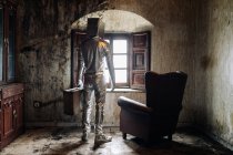 Back view unrecognizable person wearing protective silver suit with box on head standing near armchair in shabby room in abandoned house — Stock Photo