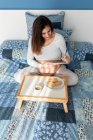 Зверху вагітна жінка сидить на ліжку і пише в блокноті під час сніданку вранці — стокове фото