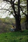 Seitenansicht einer Reisenden mit Reiseführer aus Papier, die auf einer Wiese zwischen grünen Bäumen sitzt und in der Landschaft montiert ist — Stockfoto