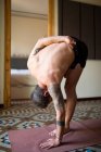Vista lateral do macho sem camisa em pé em Ardha Baddha Padmottanasana no tapete enquanto equilibra e pratica ioga em casa — Fotografia de Stock