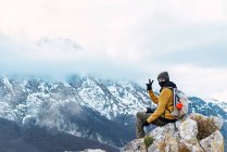 Vista lateral del excursionista con mochila mostrando dos dedos mientras está sentado en el punto alto de increíble cresta de Picos de Europa en las nubes - foto de stock