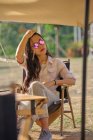 Belle femme asiatique ethnique dans des lunettes de soleil assis à la table tout en ayant un moment de détente dans la zone de camping pendant les vacances en regardant loin — Photo de stock