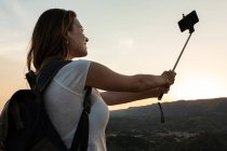 De baixo viajando fêmea com mochila em pé na colina e tomando auto-tiro no smartphone no fundo da gama de montanhas no verão — Fotografia de Stock