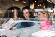 Through glass of cheerful young multiracial female best friends in trendy outfits smiling and showing two fingers gesture while taking selfie on smartphone standing near window — Stock Photo