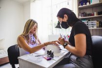 Side view ethnic female master in sterile mask applying yellow polish on nails of anonymous client in beauty salon — Stock Photo