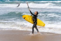 Atleta feminina em roupa de mergulho com barra de controle olhando para longe na costa arenosa contra o oceano espumoso depois de praticar kiteboarding — Fotografia de Stock