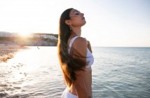 Seitenansicht einer jungen Frau in weißer Badebekleidung und Ohrring, die gegen welliges Meer mit Halterung wegschaut — Stockfoto