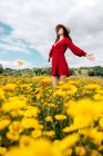 Снизу вид на модную женщину в красном сарафане и с цветочной короной, стоящей с закрытыми глазами на цветущем поле с желтыми и красными цветами с протянутыми руками в теплый летний день — стоковое фото