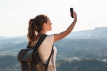 Voltar ver feliz jovem caminhante feminino com mochila tirando selfie no smartphone enquanto está em pé no topo da colina verdejante — Fotografia de Stock