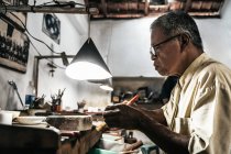 Vue latérale d'un homme ethnique mature travaillant à un bureau endommagé dans un atelier artisanal — Photo de stock