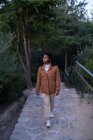 Giovane donna afroamericana calma in giacca calda in piedi nel verdeggiante parco lussureggiante e guardando lontano nella giornata primaverile — Foto stock
