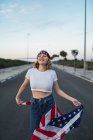 Encantada mulher americana de pé com bandeira nacional dos EUA na estrada ao pôr-do-sol e olhando para cima — Fotografia de Stock