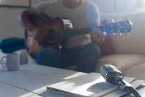 Vintage microphone placed on table near crop unrecognizable bearded male musician playing acoustic guitar on sofa — Stock Photo