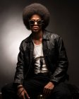Fashionable African American male in leather jacket and boots sitting on gray background — Stock Photo