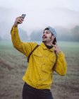 Sac à dos souriant jeune homme en jean jaune prenant selfie sur smartphone tout en se tenant debout sur la prairie brumeuse dans la nature — Photo de stock