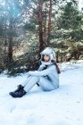 Side view of focused young female astronaut in spacesuit and helmet looking away and sitting in snowy woodland — Stock Photo