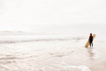 Vista posterior de un surfista irreconocible vestido con traje de neopreno de pie mirando hacia el agua con tabla de surf para atrapar una ola en la playa durante el amanecer - foto de stock