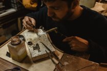 Бородатий чоловічий золотошукач використовує щипці, роблячи крихітні металеві деталі на верстаті в майстерні — стокове фото