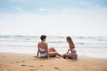 Vue arrière de jeunes amies souriantes méconnaissables en maillots de bain blancs assis sur un rivage sablonneux près de l'océan sous un ciel nuageux bleu par temps ensoleillé — Photo de stock