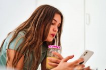 Vue latérale de la jeune étudiante naviguant sur les réseaux sociaux sur un téléphone portable près de la table avec des fruits frais et du jus tout en passant la matinée à la maison — Photo de stock