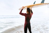 Vista lateral do jovem surfista irreconhecível com cabelo comprido vestido de fato de mergulho em pé olhando para a praia com a prancha na cabeça durante o nascer do sol — Fotografia de Stock