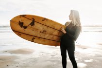 Жінка-серфер, одягнена в гідрокостюм, стоїть озираючись з дошкою для серфінгу на пляжі під час сходу сонця на задньому плані — стокове фото