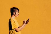 Vista laterale della donna contemporanea con elegante taglio di capelli e piercing utilizzando lo smartphone per inviare messaggi di testo sui social media su sfondo giallo — Foto stock