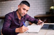Привабливий молодий іспаномовний чоловік - підприємець перевіряє інформацію на смартфоні і пише нотатки в планувальнику, працюючи за столом з ноутбуком. — стокове фото