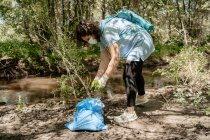 Активна жіноча екологічна волонтерка в захисній масці і рукавичках, що збирають сміття з струмка в мішок для сміття в літньому лісі — стокове фото