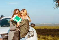 Fröhliche junge Freundinnen mit Low-Gang-Schild stehen an weißem Auto am Ufer des Meeres und umarmen sich beim Trip — Stockfoto