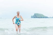 Hembra envejecida en forma activa positiva en traje de baño caminando fuera del agua de mar mientras disfruta del día de verano en la playa - foto de stock