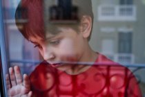 Através de vidro de menino pré-adolescente infeliz com hematomas no rosto olhando para longe enquanto estava perto da janela em casa como conceito de violência doméstica e abuso infantil — Fotografia de Stock