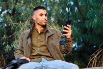 Hispanic guy in casual clothes looking away and texting on phone while standing near barrier and coniferous tree in park — Stock Photo