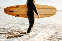 Вид збоку обрізаний невпізнаваний серфінг чоловік, одягнений в гідрокостюм, стоїть з дошкою для серфінгу на воді, чекаючи, щоб спіймати хвилю на пляжі під час сходу сонця — стокове фото