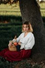 Junge Frau in altmodischer weißer Bluse und Rock hält Weidenkorb voller frischer Äpfel in der Hand und blickt in die Kamera, während sie sich an einem Sommertag in der Natur am Baum ausruht — Stockfoto