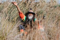 Alegre rastafari étnico de edad con rastas mirando a la cámara en pie en un prado seco en la naturaleza - foto de stock