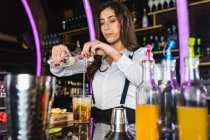 Женщина-бармен в стильном наряде добавляет жидкость из бутылки в стекло и перемешивает длинной ложкой, готовясь коктейль стоя у стойки в современном баре — стоковое фото