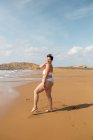 Full length of young female in swimsuit standing looking at camera on sandy coast in sunny day under blue cloudy sky — Stock Photo