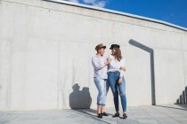 Полное тело молодых положительных женщин-друзей в модных нарядах и шляпах, стоящих, глядя друг на друга на дорожке рядом с серой стеной в солнечный день под голубым небом — стоковое фото