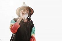 Fröhlicher alter Rastafari mit Dreadlocks, der in die Kamera schaut, während er auf einer trockenen Wiese in der Natur Unkraut raucht — Stockfoto