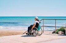 Vista laterale viaggiatore femminile in sedia a rotelle con zaino godendo il viaggio estivo sulla spiaggia vicino al mare blu — Foto stock