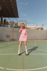 Piena lunghezza gioiosa vestibilità femminile in rosa prendisole pattinaggio su rulli scattare foto sulla fotocamera istantanea sul terreno sportivo soleggiato — Foto stock
