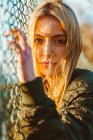 Belle femme blonde en veste regardant la caméra debout à la clôture de maillon de chaîne en lumière du soleil dorée — Photo de stock